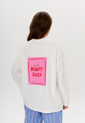Witte oversized sweatshirt met een roze rechthoekige graphic op de achterkant met de tekst "Ik heb mijn SCHOONHEIDSSLAAP nodig" in vetrode letters.