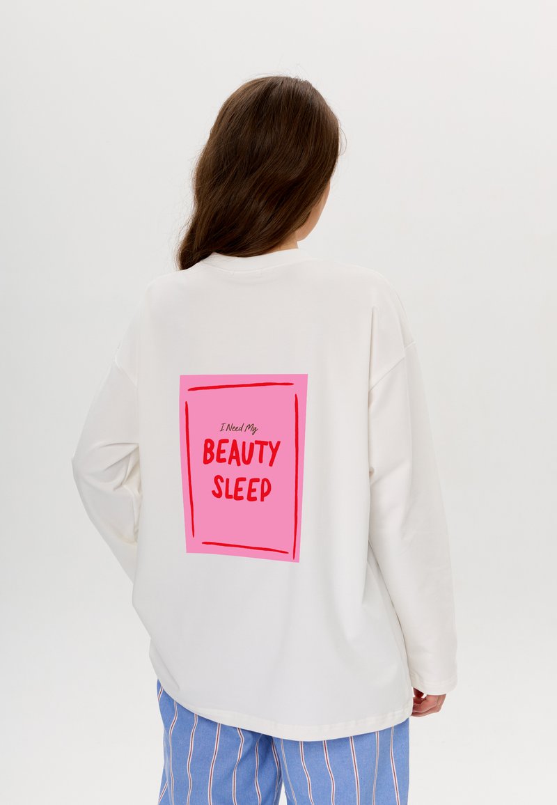 Weißes, übergroßes Sweatshirt mit einem pinken rechteckigen Grafikdruck auf dem Rücken, der den Text "Ich brauche meinen Schönheitsschlaf" in fetten roten Buchstaben zeigt.