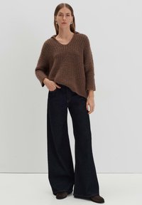 Brauner gestrickter Pullover mit V-Ausschnitt, der ein strukturiertes Muster aufweist. Kombiniert mit weit geschnittenen, dunkelblauen Jeans und flachen braunen Schuhen.