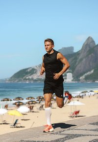 Biegaczka nosi czarną bezrękawnik i szorty, a na nogach ma białe skarpety kompresyjne oraz jaskrawo pomarańczowe buty, na promenadzie wzdłuż plaży.