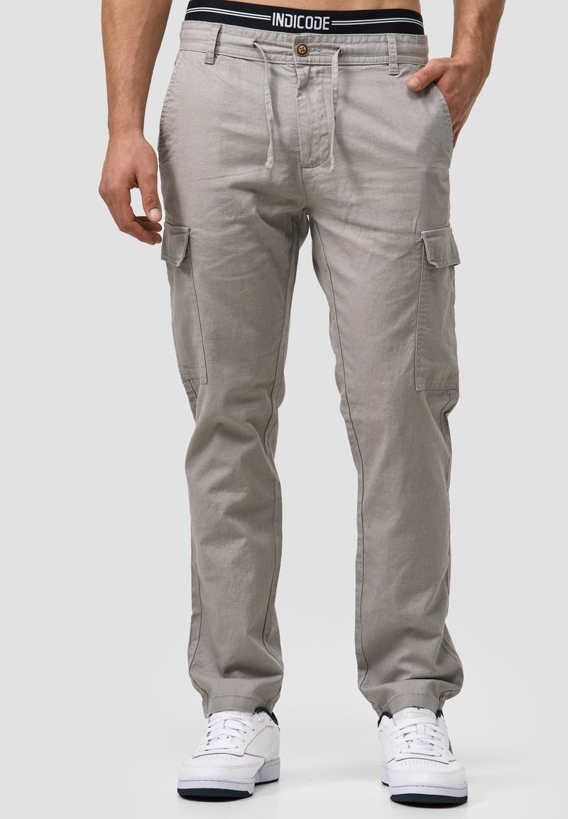 Helle graue Cargo-Hosen mit sichtbaren Nähten, mehreren Seitentaschen und einem elastischen Bund mit schwarzem Logo. Kombiniert mit weißen Sneakers.