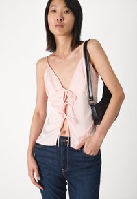 Top de tirantes finos en rosa claro con escote en V profundo y detalle de nudo frontal. Combinado con jeans de mezclilla oscuros y un bolso negro. Textura suave.