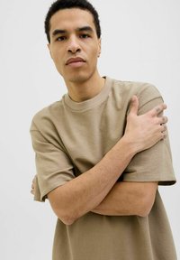 Beige kortærmet t-shirt med rund hals, afslappet pasform og struktureret stof. Modellen krydser armene og viser subtile tatoveringer på hænderne.