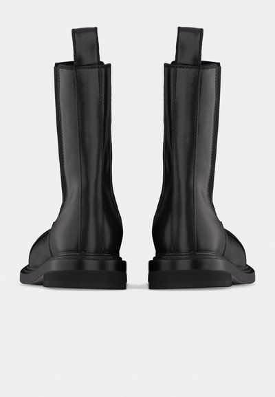 Ζευγάρι μαύρες δερμάτινες μπότες μεσαίου μήκους μέχρι τον αστράγαλο, με θηλιές τράβηγματος, εμφανή ραφές και επίπεδες λαστιχένιες σόλες, εμφανιζόμενες από πίσω σε λευκό φόντο.