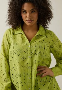 Camicia in cotone verde con colletto, caratterizzata da ricami a occhiello e motivi floreali, maniche lunghe e chiusura frontale con bottoni.