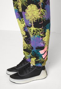 Black sneakers with white soles worn with black pants featuring colorful tree and root patterns and pink "Stella McCartney" text.