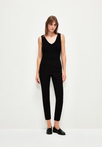 Zwarte mouwloze top met een V-hals, gecombineerd met op maat gemaakte zwarte broek. Platte zwarte schoenen maken de outfit compleet en benadrukken een strakke uitstraling.