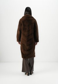 Donna di spalle con indosso un lungo cappotto di pelliccia marrone scuro sopra pantaloni grigi a gamba larga e scarpe nere con tacco, su uno sfondo bianco semplice.