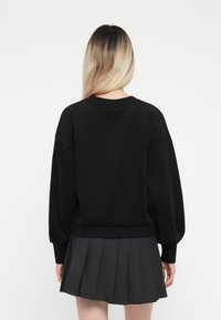 Svart oversized sweatshirt med lange, puffede ermer og ribbestrikket kant, kombinert med et plisserte mørkegrå skjørt. Glatt stoff tekstur.