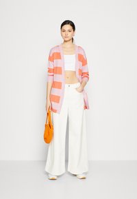 Pruhovaný růžový a oranžový cardigan přes bílý crop top, doplněný širokými bílými kalhotami. Drží texturovanou oranžovou tašku. Tenisky dokončují outfit.