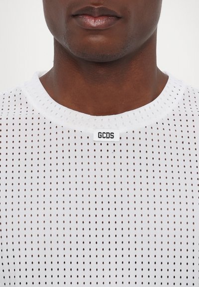 T-shirt bianco in rete con perforazioni circolari, dotato di un collo rotondo e un'etichetta con "GCDS" vicino al colletto. Texture liscia.