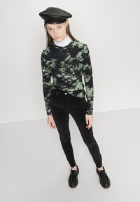 Top de manga longa preto e verde com efeito tie-dye, combinado com calças de veludo pretas e sapatos pretos, apresentando um chapéu preto brilhante.