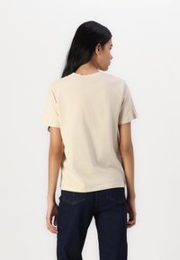 Beige bomull t-skjorte med rund hals og korte ermer, vist fra baksiden. Kombinert med mørkeblå denimjeans.