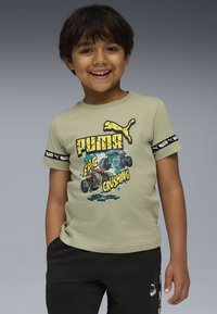 Lächelnder Junge in einem hellgrünen Puma-T-Shirt mit Monstertruck-Motiv und schwarzen Hosen mit Puma-Logo vor einem grauen Hintergrund.