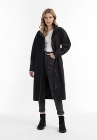 Zwarte lange jas met een hoge kraag, ritssluiting aan de voorkant en drukknoopjes. Draagt over een wit overhemd en donkere jeans, gecombineerd met zwarte veterschoenen.