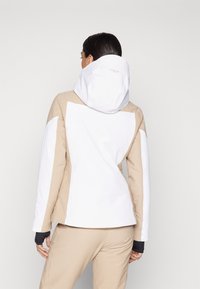 Skijacke in Beige und Weiß, mit Kapuze, tailliertem Design, strukturiertem Stoff an den Ärmeln und einem Logo am Kragen.