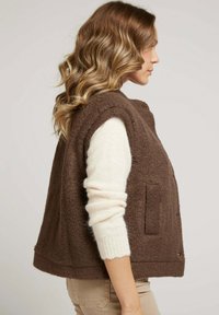 Gilet en polaire marron au design décontracté, avec un col haut et des poches latérales, superposé sur un pull crème, vu de côté.