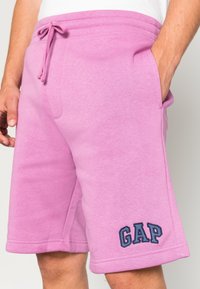 Pantalones cortos de algodón rosa con cintura ajustable, bolsillos laterales y un logo bordado de "GAP" en color marino en la pierna inferior. Textura suave.