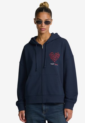 Femme avec des lunettes de soleil portant un sweat à capuche bleu marine zippé avec un motif cœur rouge et le texte "SWEET heart" sur la poitrine gauche, associé à un jean bleu.