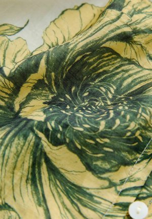 Auduma ar ziedu rakstu zaļās un dzeltenās toņos, ar maigu tekstūru un ziedu kontūrām. Ietver balta pogas detaļu.