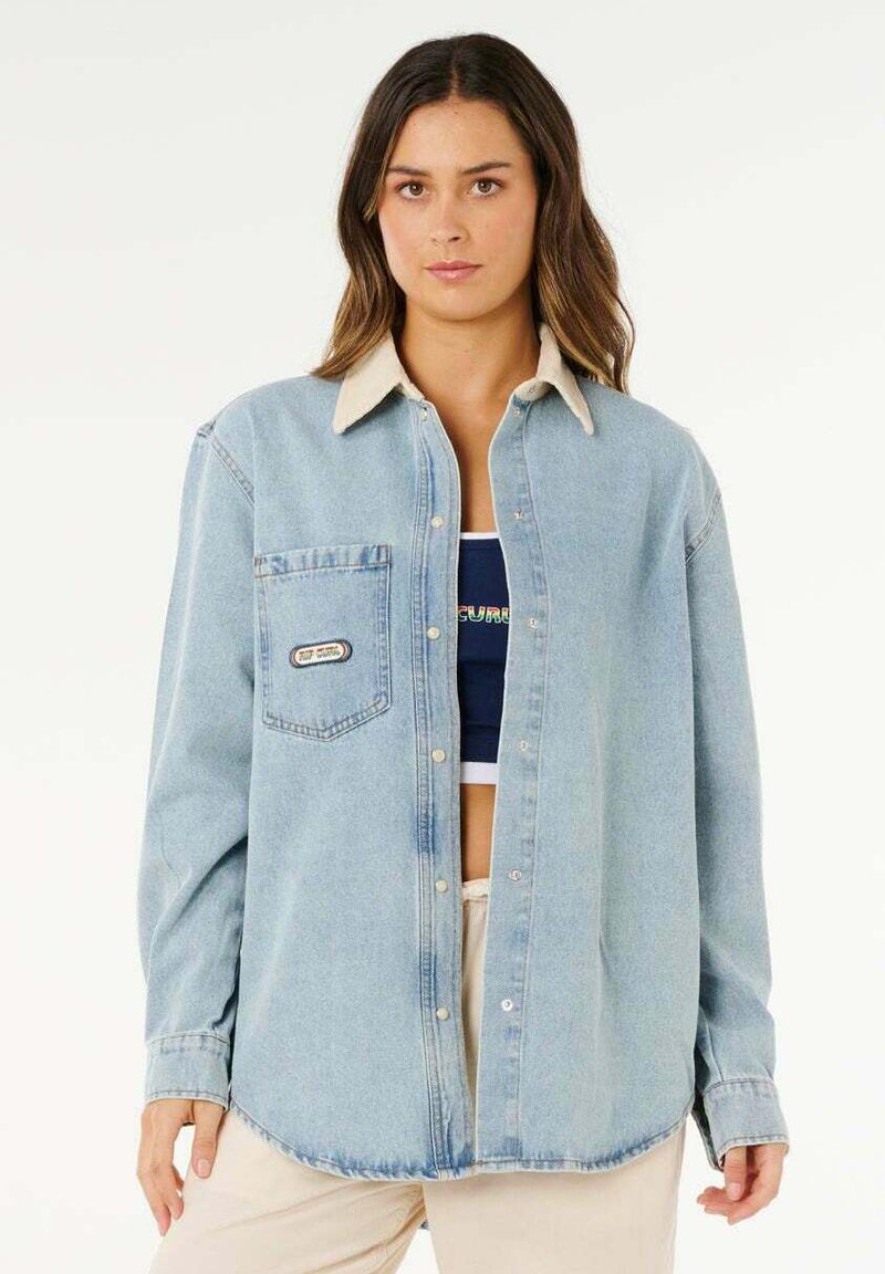 Frau mit langem braunem Haar, die ein offenes hellblaues Jeanshemd über einem schwarzen Crop-Top und beigen Hosen trägt, steht vor einem weißen Hintergrund.