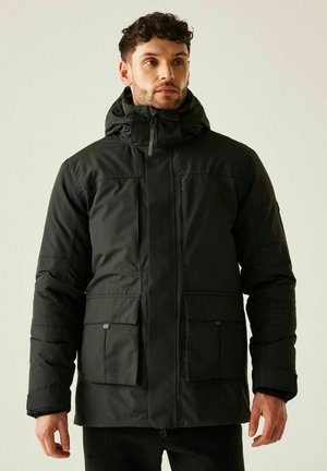 Mężczyzna w czarnej ocieplanej zimowej kurtce z kapturem i przednimi kieszeniami, stojący na tle jasnego, jednolitego tła.