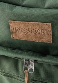 Πράσινο σακίδιο Jansport με καφέ σουέντ ετικέτα λογότυπου και δύο μεταλλικούς φερμουάρ με χαραγμένο "JS" στην μπροστινή τσέπη.