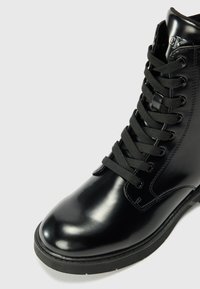 Bottine montante en cuir noir brillant à lacets, semelle épaisse et œillets en métal, avec logo CK embossé près du haut.