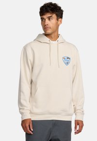 Beige hoodie met een voorzak, een kap met trekkoord en een kleurrijk logo op de linkerkant van de borst. Zacht materiaal en een relaxed fit.