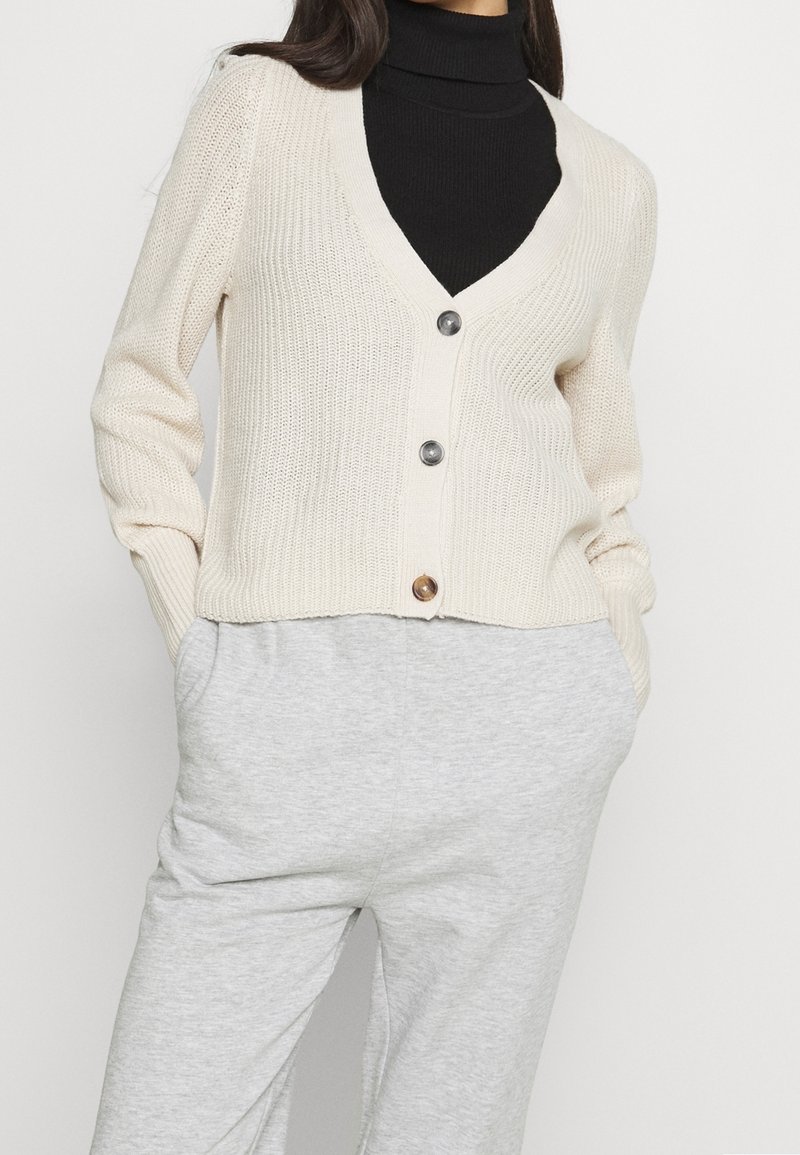Femme portant un cardigan en maille crème à trois boutons par-dessus un col roulé noir et un pantalon gris clair, les mains dans les poches.