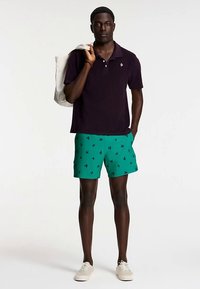 Polo majica tamnoljubičaste boje, mješavina pamuka; zelene kupaće hlače s crnim uzorkom ptica; bež tenisice; drži svijetlu torbu za nošenje.