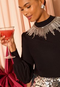 Pullover nero con maniche a sbuffo, adornato con un colletto di paillettes argento. La modella tiene un bicchiere da martini trasparente con una bevanda rosa.