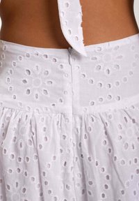 Vestido de algodón blanco con un patrón de ojales florales bordados, cierre en la parte trasera con cremallera y detalle de tiras atadas en el escote. Tejido texturizado.