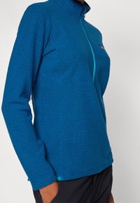 Modrý fleece pulovr s vysokým límcem, texturovaným vzorem, zipovým zapínáním a kontrastními tyrkysovými detaily na lemu.