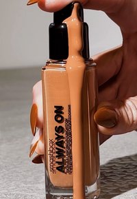 Flacon de fond de teint Smashbox Always On avec une base en verre transparent, un pompe noire, et un liquide moyen tan s'écoulant du bec, tenu par une main.