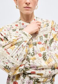 Γυναίκα που φοράει μπλούζα με λουλουδάτο σχέδιο, λεπτομέρειες δαντέλας και φουσκωτά μανίκια, κρατώντας το γιακά κοντά στο πηγούνι της, με ορατά χρυσά σκουλαρίκια κρίκους.