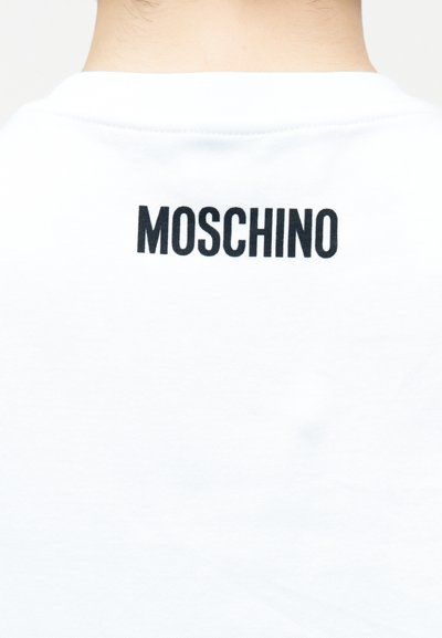 Πίσω μέρος μιας λευκής μπλούζας με μαύρο κείμενο "MOSCHINO" τυπωμένο κοντά στον γιακά στον λαιμό και το πάνω μέρος της πλάτης ενός ατόμου.
