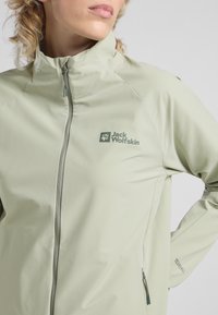 Giacca verde chiaro con zip realizzata in tessuto liscio. Presenta il logo Jack Wolfskin, colletto alto e tasche con zip. Design semplice senza motivi.