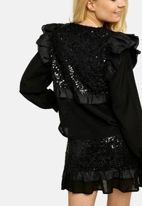 Melnas volānu garām piedurknēm tops ar spīguļu akcentiem un atbilstoša spīguļu svārka ar volānu apakšu. Teksturēts audums ar spīdīgiem elementiem.