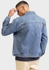 Giacca di jeans blu chiaro con taglio classico, cuciture visibili e due tasche laterali. Presenta un colletto e un dettaglio con una toppa sul retro.