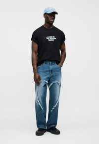 Mladý muž nosiaci čierne tričko s vyhrnutými rukávmi a bielym nápisom, roztrhané modré džínsy s opotrebovanými náplasťami v tvare srdca, čierne topánky a svetlomodrá čiapka.