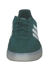 Zelené semišové tenisky s bílými pruhy, šněrovacím designem, polstrovaným límcem a průhlednou podešví. Pohled zepředu zvýrazňuje detaily svršku.