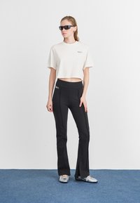 Femeie îmbrăcată cu pantaloni negri cu evazare și fante laterale, un tricou alb scurt, pantofi plat din argint și ochelari de soare negri, stând pe un covor albastru.