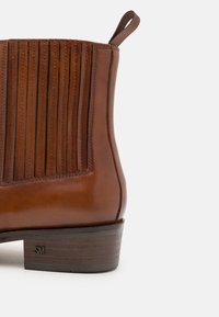 Botín de piel marrón hasta el tobillo con canalones verticales, punta redondeada y pequeño tirador. Tacón de madera resistente con el logo "SM" estampado.