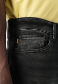 Jeans din denim negri cu o textură estompată, având un etichetă de buzunar inscripționată "BOSS". O cămașă galbenă este parțial vizibilă deasupra jeanșilor.