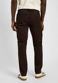 Brązowe bawełniane spodnie o prostym kroju, z dwoma tylnymi kieszeniami i gładką fakturą. Nosi się je z białymi butami z ciemnymi paskami.