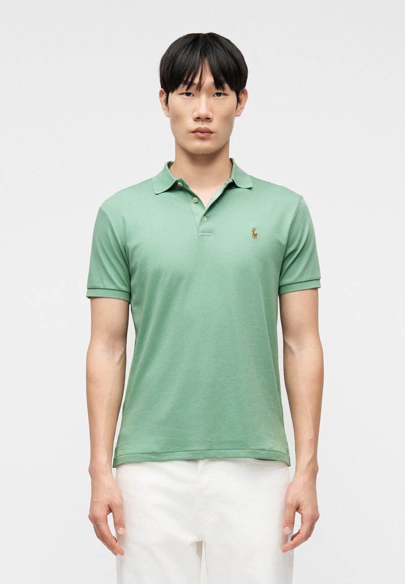 Νεαρός άνδρας με ανοιχτό πράσινο π όλο και λευκό παντελόνι, στέκεται μπροστά σε απλό λευκό φόντο, κοιτάζοντας προς τα εμπρός.