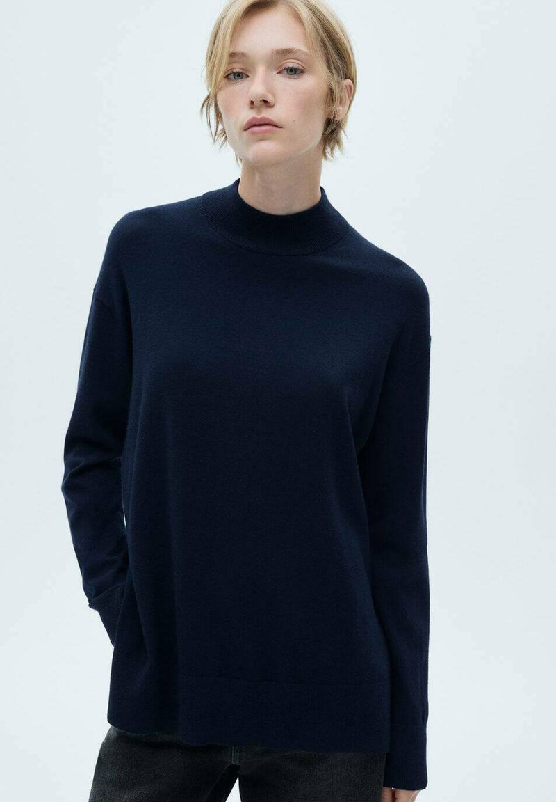 Pulover bleumarin cu guler mock turtleneck, material tricotat moale, croi lejer, mâneci lungi și tiv cu margine ondulată. Asortat cu blugi închiși la culoare.
