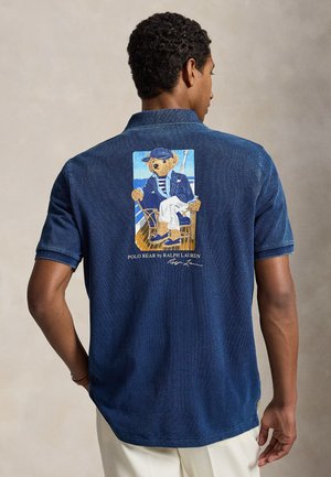 Homme portant un polo bleu marine avec un motif coloré représentant un ours vêtu d'une tenue de marin assis sur une chaise au bord de la mer.