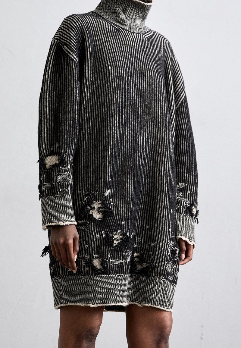 Robe en tricot rayée noir et blanc à manches longues avec col roulé, comportant plusieurs trous intentionnels et des détails usés sur les manches et l'ourlet.